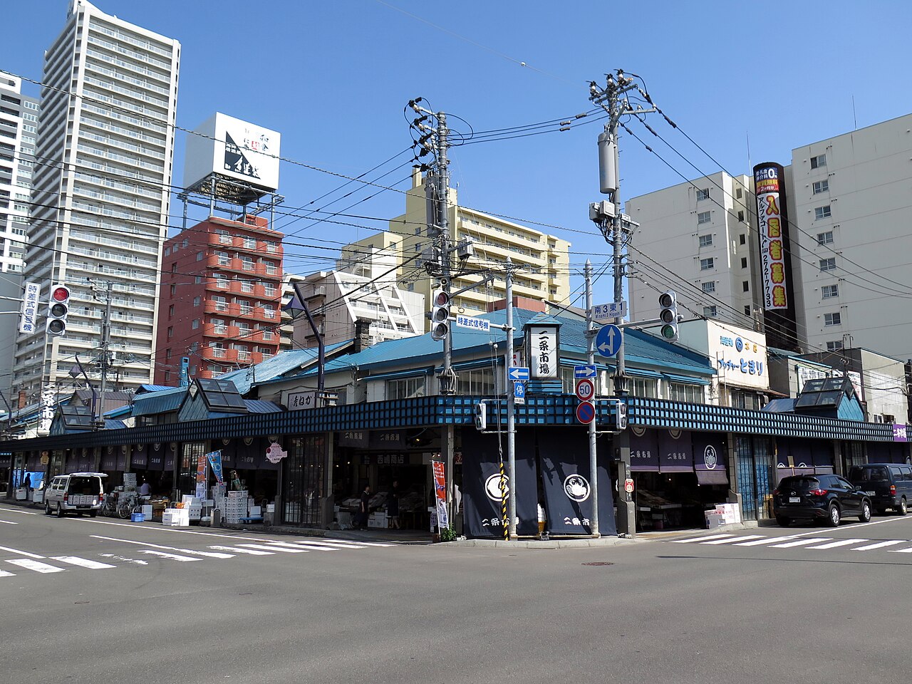 札幌・小樽2泊3日モデルプラン|絶品海鮮とレトロ街歩きを満喫する旅 2 交差点から見た二条市場の外観。青いトタン屋根の平屋が連なり、縦看板と紺色の暖簾が出ている