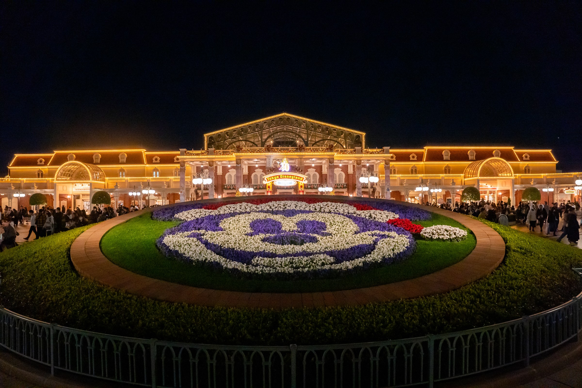 夜の東京ディズニーランド、メインエントランス前のミッキーの花壇とライトアップされた駅舎風の建物