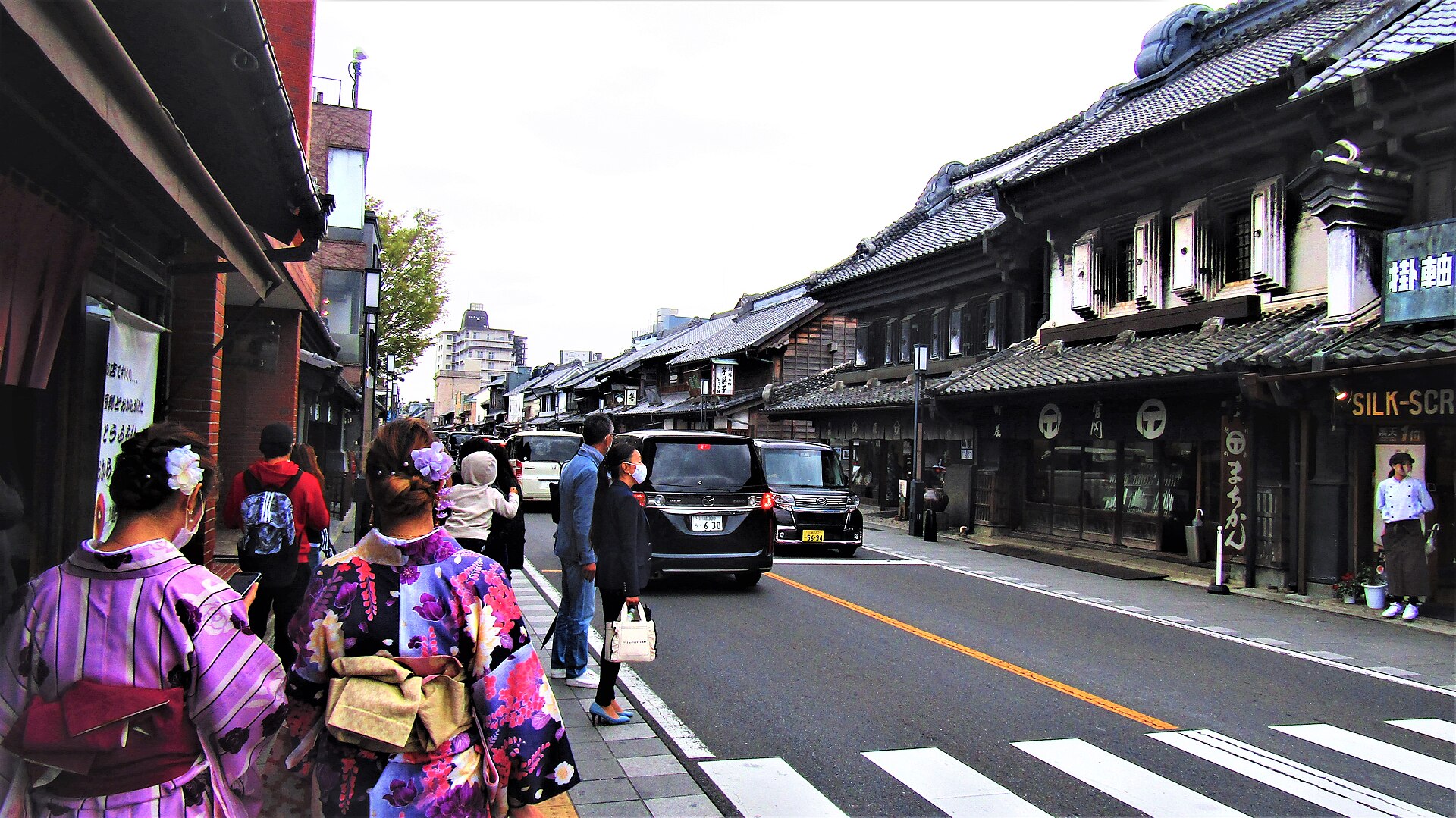 川越の蔵造りの町並み。黒漆喰の店蔵が通りの片側に連なり、着物姿の人が歩道を歩いている