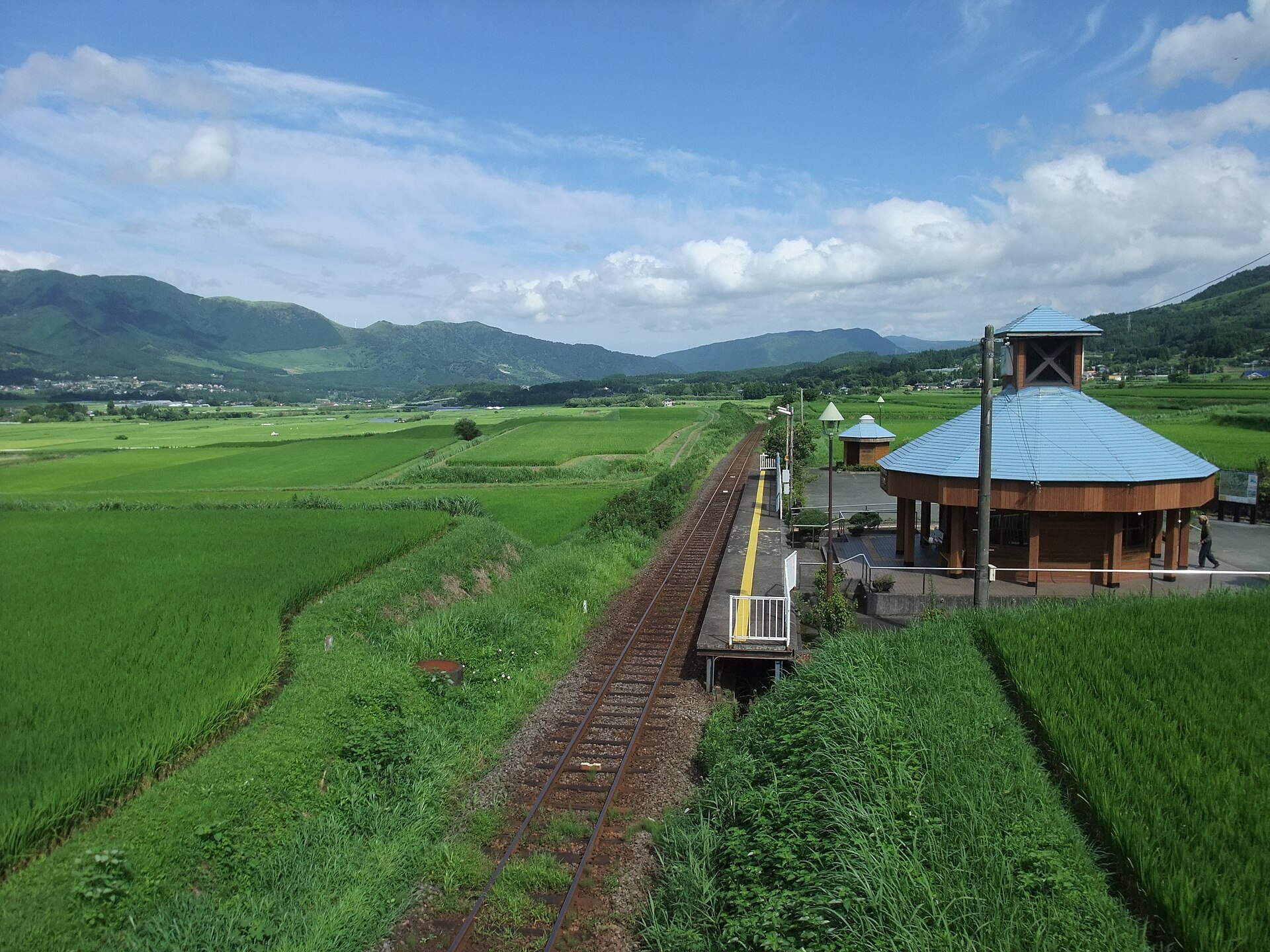 TAOYA阿蘇の料金|オールインクルーシブに含まれるものと別料金 1 南阿蘇鉄道の南阿蘇水の生まれる里白水高原駅。線路と水田の向こうに山並みが見える
